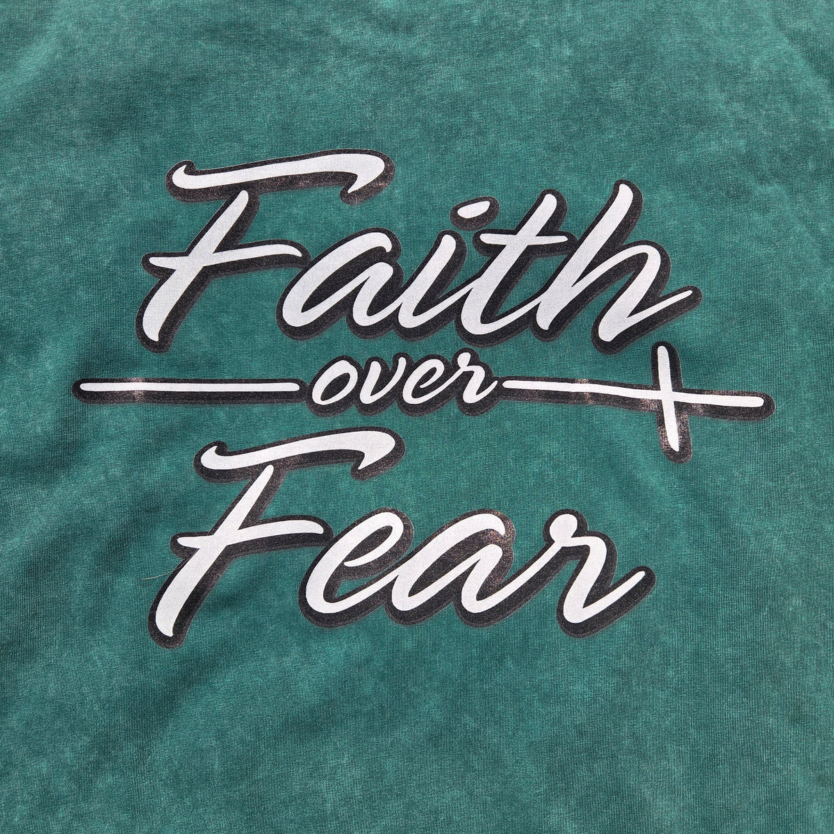 A close-up of a teal-green mineral-wash t-shirt featuring the phrase 'Faith over Fear' printed in white script with a dark outline across the chest.